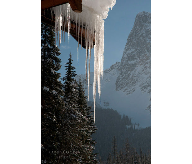 Detail of thick, clear icicles on the gable of a roof, overlooking a steep, snow-capped mountain and large, snow covered evergreen tees. The sun is shining on the ice. 