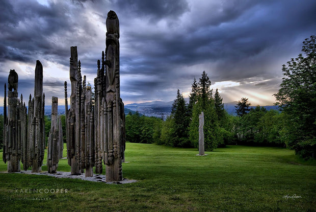 Fine art luxury nature landscape contemporary photography by Karen Cooper Gallery Various Ainu wooden sculptures in a green park on Burnaby Mountain, overlooking the storm-covered sky with sun rays jutting through a small hole onto the mountains and city in the background Vancouver British Columbia Canada