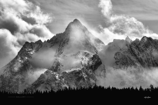 Black and white of one of the Seven Sisters mountain peak. Base of mountain dominated by the profile of evergreen trees. Rugged sharp peaks covered with rolling, thin clouds. 