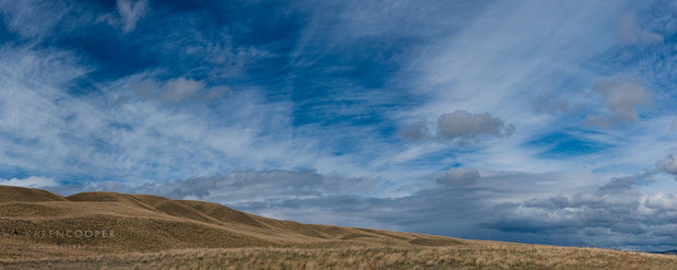 Rolling hills covered in yellow patches of grass. Large, blue sky covered in wisping cloud trails and patches of grey clouds. 
