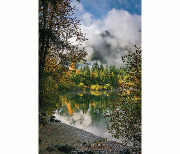 Lush green and yellow autumn trees overlooking a rocky beach with a still lake and a cloud-covered mountain