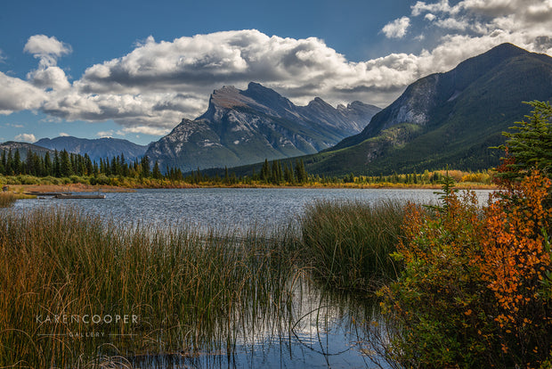 Vermillion Lake Alberta with rugged Mount Rundle in Background against blue, cloud-filled sky, surrounded by lush colourful fall foliage. 
