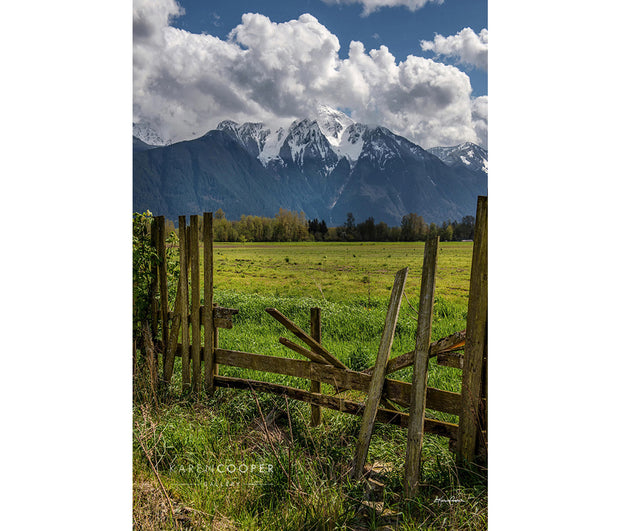 Broken wooden fence in foreground, with a green pasture and trees in the distance. Background filled by large, snowy mountain peak partially covered with thick white clouds and a Blue sky. 