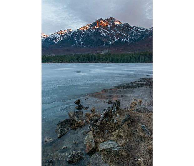 A frozen lake and shoreline with yellow grass, tree branches, and rocks. Overlooking the rugged mountain with veins of snow, its peaks orange from the morning sun. 