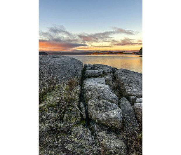 A yellow and red cloud-filled sky reflecting in the ocean in the background. Foreground dominated by a large, grey rocky cliff. 