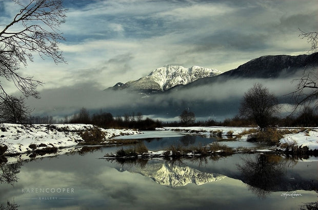 A snow covered meadow shadowed by a snow-capped mountain. The stream below is perfectly reflecting the mountain and trees