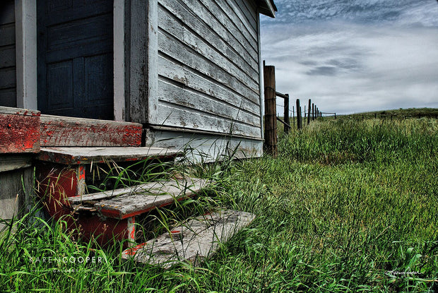 Detail of weathered stairs leading up to the porch of an old, abandoned farmhouse, surrounded by clouds, a barbed wire fence, and short green grasses 