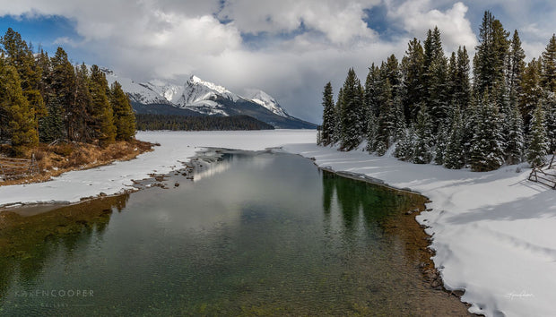 A clear, shallow river with their shores covered in white, pristine snow. Flanked by two sets of evergreen trees, and in the centre background a tall, jagged, snow covered mountain framed by thick white clouds with patches of blue sky. 
