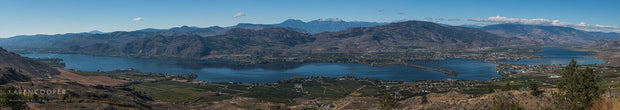 A wide panorama overlooking a large lake, surrounded by large rolling hills that frame the valley. landscape dominated by fields, houses, and trees. 